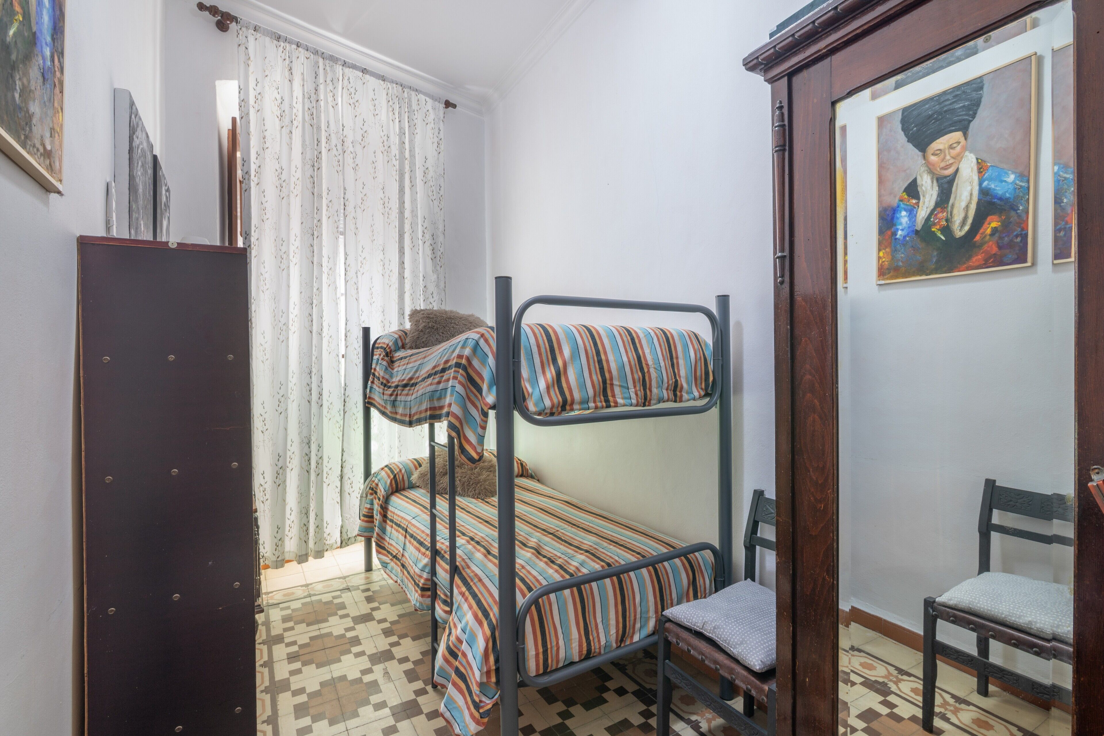 Une chambre avec un lit superposé, une couvre-lit à motifs, une armoire en bois, une chaise et un tableau accroché au mur.