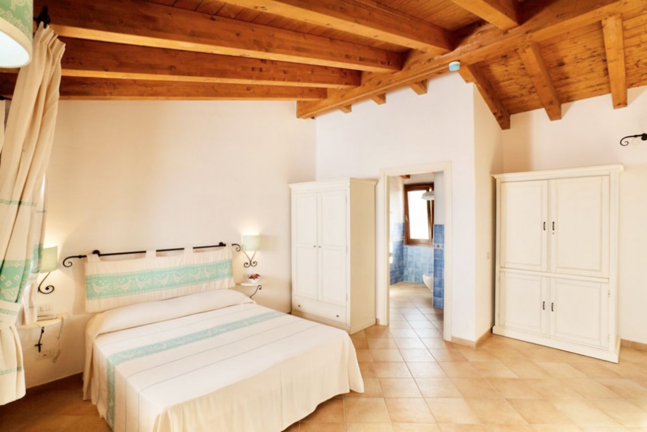 Une chambre avec un plafond en bois, un lit recouvert d’une courtepointe à rayures, des armoires blanches et une salle de bain visible à travers une porte ouverte.