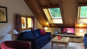 A cozy living room with a blue sofa, red chair, wooden coffee table, and a TV on a wooden stand. - Chalet in Huez near Ski Slope (Oz en Oisans)