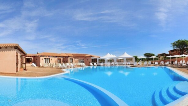 Una gran piscina con sillones de playa y sombrillas, rodeada de edificios de estilo mediterráneo.