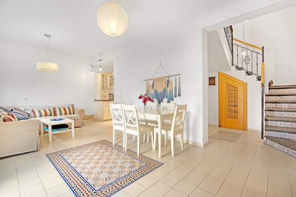 A living room with a sofa, dining table, chairs, a patterned rug, a hanging light fixture, and a staircase.