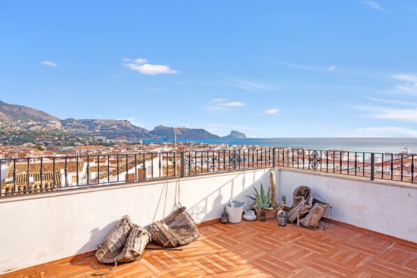 A rooftop terrace with a view of the city and sea, wooden flooring, and some potted plants.