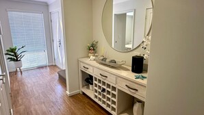 Un baño con un espejo grande, un lavamanos con un lavabo decorativo, una planta en maceta y una estantería para vinos.