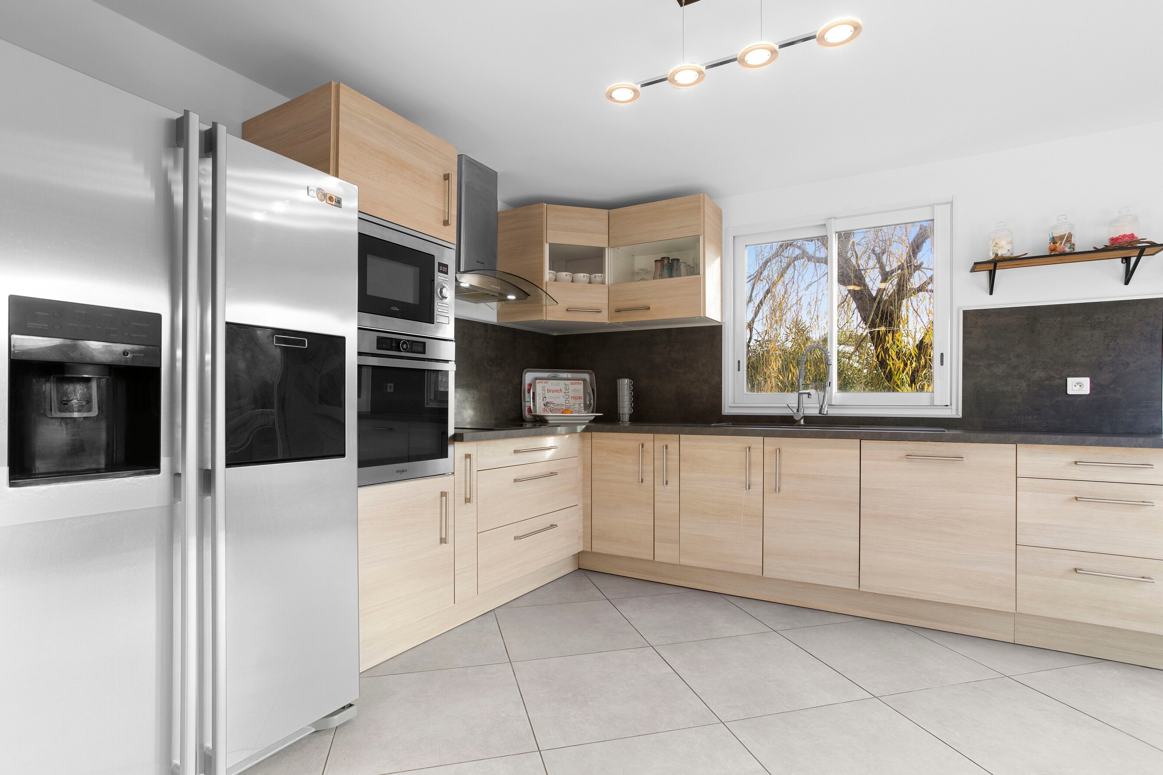A modern kitchen with a stainless steel refrigerator, built-in oven, microwave, and a window with a view of trees.