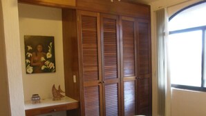 A bedroom with a bed, a wooden wardrobe, a small shelf with decorative items, and a window with curtains.