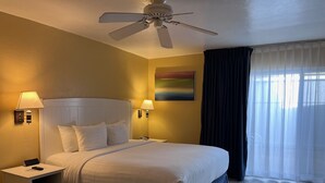 Une chambre d’hôtel avec un lit, deux lampes de chevet, un ventilateur de plafond, un tableau au mur et une fenêtre avec des rideaux.