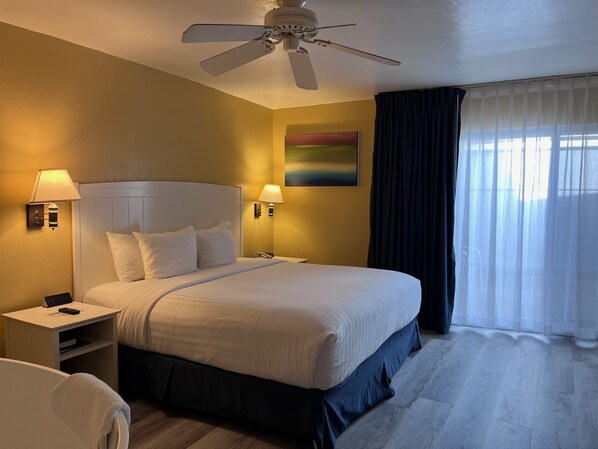 Une chambre d’hôtel avec un lit, deux lampes de chevet, un ventilateur de plafond, un tableau au mur et une fenêtre avec des rideaux.