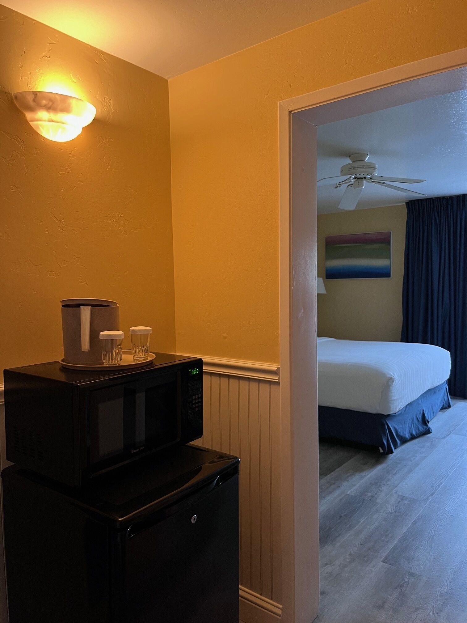 Une chambre d’hôtel compacte comprenant un coin cuisine, un lit, un ventilateur de plafond et un tableau au mur.