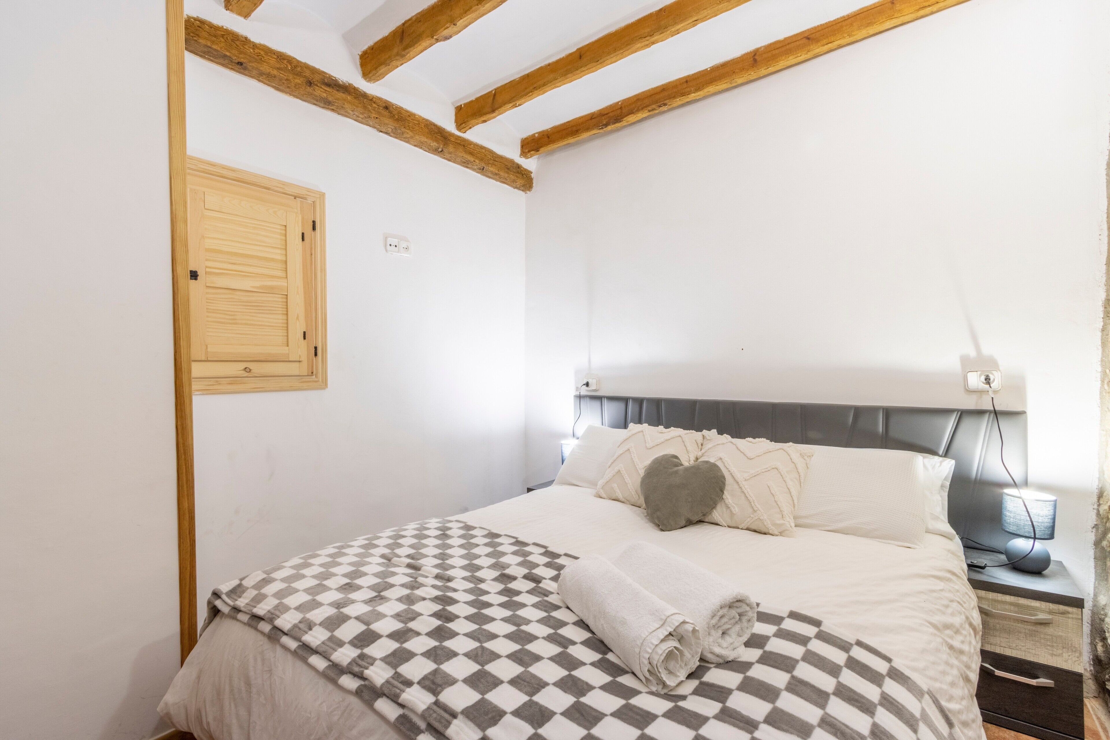 Quarto com teto de madeira, cama com manta xadrez, duas almofadas, mesa de cabeceira com abajur e interruptor de luz na parede.