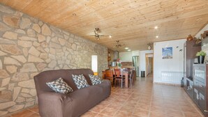 Una sala de estar con pared de piedra, techo de madera, un sofá marrón con cojines estampados, un comedor con mesa y sillas, y una zona de cocina con armarios y un refrigerador.