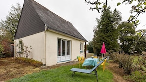 Une petite maison avec un toit en pente et une grande fenêtre. On y trouve une pelouse avec une table et des chaises, ainsi qu’un parasol rose.