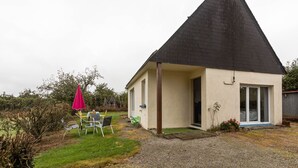 Une petite maison avec un toit en pente, une allée gravillonnée et une cour verdoyante agrémentée d’un parasol rouge et de mobilier d’extérieur.