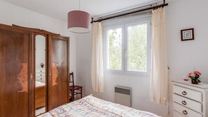 Une chambre à coucher comprenant un lit, une armoire, une chaise, une commode, une fenêtre avec des rideaux et un luminaire suspendu.