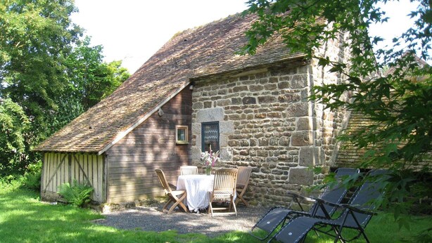 A rustic stone building with a sloped roof, a small table with chairs, and a garden area with a lawn and a tree. - Holiday Home in Ceaucé near Mont Saint Michel (Céaucé)