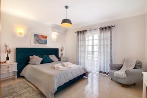 Une chambre à coucher comprenant un lit, une chaise, une table de chevet, une lampe, un tableau et une fenêtre avec des rideaux.