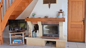 A cozy living room with a fireplace, wooden staircase, and a television on a stand. - Holiday Home in Vosges with Garden and BBQ (Le Haut-Du-Them)