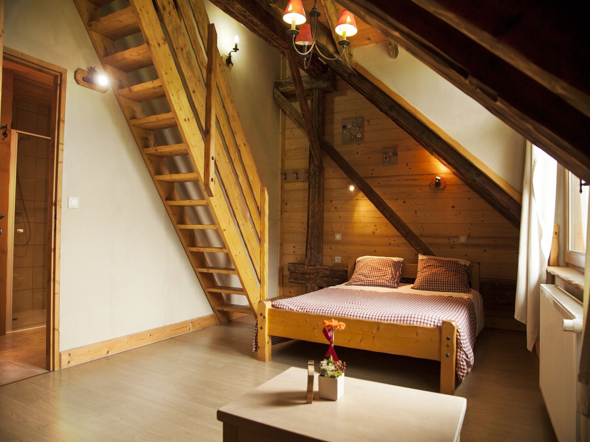A cozy attic bedroom with a wooden staircase, a bed with checkered bedding, and a small table with a flower vase.