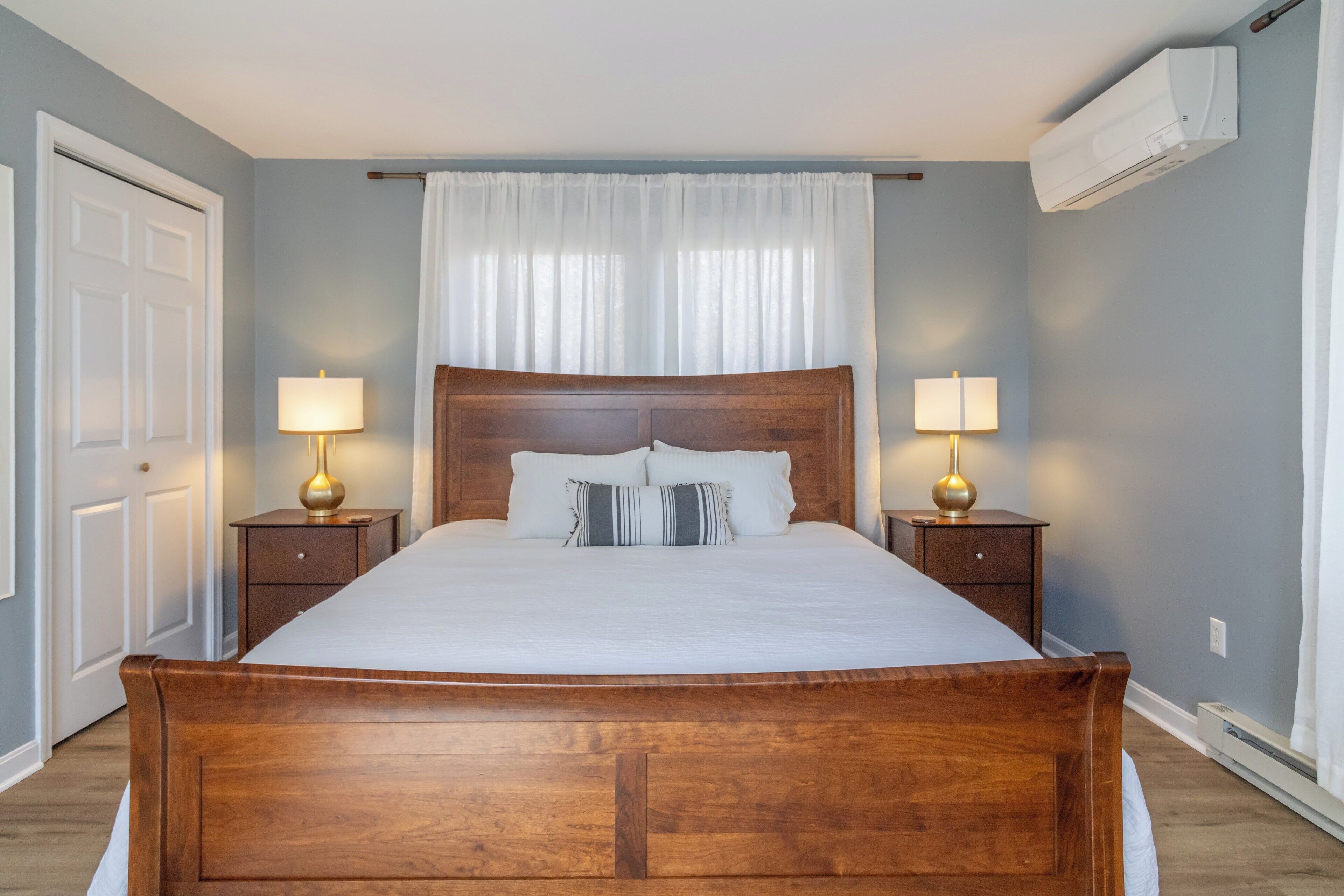 A bedroom with a wooden bed, two bedside tables with lamps, a white curtain, and an air conditioning unit.