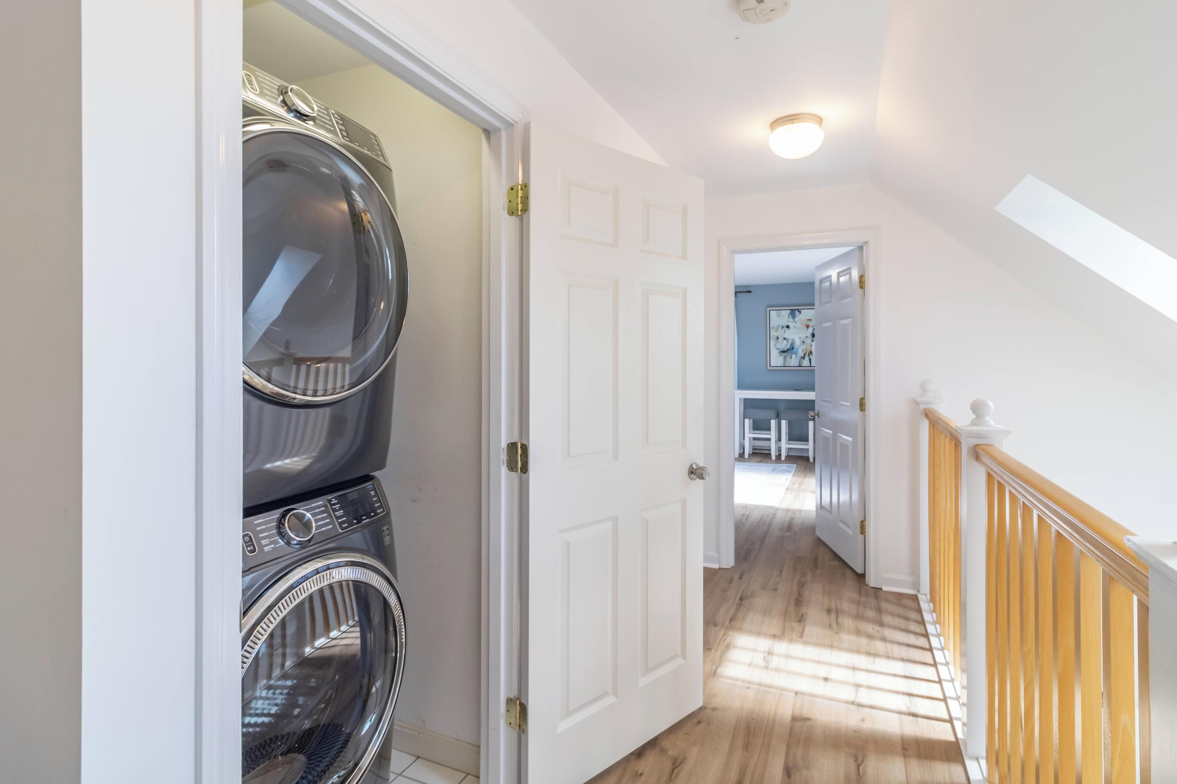 Une buanderie avec des machines à laver et des sèche-linge superposés, une porte blanche et un escalier en bois menant à une autre pièce.