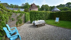 Eine gepflasterte Terrasse mit Picknicktisch und blauem Stuhl, umgeben von einer Hecke und einem Holenzaun.