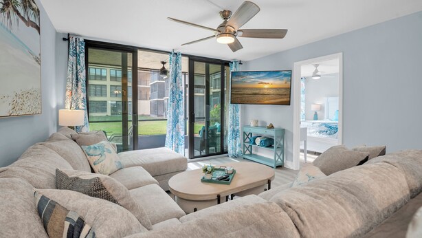A living room with a large sofa, a coffee table, and a ceiling fan. There is a sliding glass door leading to a balcony with a view of the outdoors.