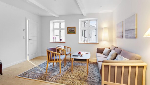 Ett vardagsrum med en soffa, två fåtöljer, ett soffbord och en matta. Det finns två fönster med gardiner, en tavla på väggen och en lampa.
