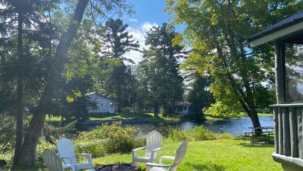 A lakeside patio with two Adirondack chairs, a fire pit, and a view of a house across the water. - Waterfront cabin with access to Long Lake and Mud Lake.   (Sinclair)