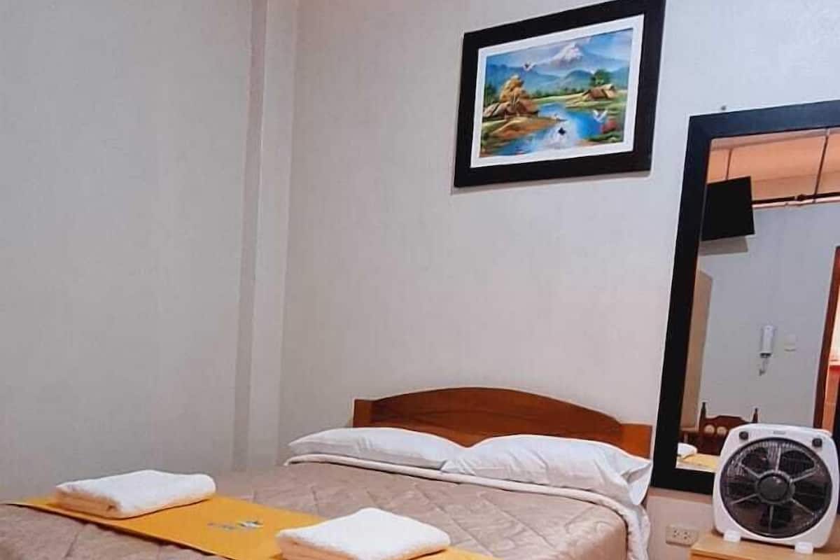A neatly made bed with a brown bedspread and two folded white towels, a small wooden nightstand with a fan, and a framed picture on the wall.