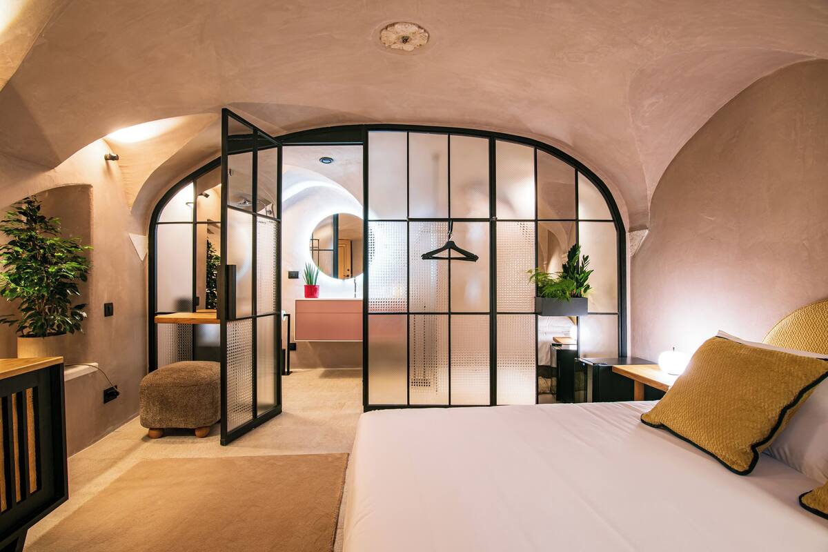 Un dormitorio con una cama grande, un banco de madera, una lámpara colgante y vista a un baño a través de un separador de vidrio.