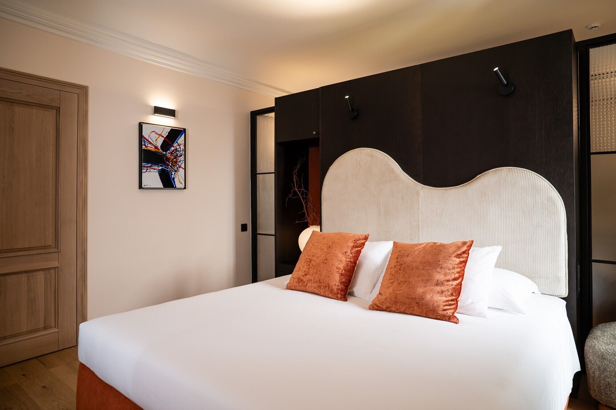 A neatly made bed with white and brown pillows, a dark headboard, and a framed abstract painting on the wall.