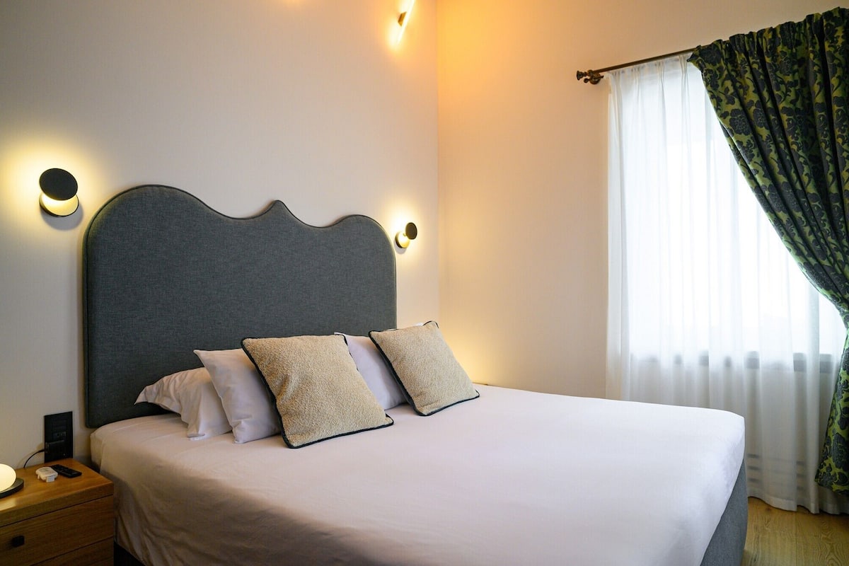 Una cama bien tendida con cabecera gris, dos almohadas blancas y una manta color beige. Hay una mesita de noche con una lámpara y un control remoto. La habitación tiene una ventana con cortinas de gasa y una cortina estampada parcialmente abierta.