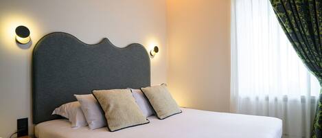 Una cama bien tendida con cabecera gris, dos almohadas blancas y una manta color beige. Hay una mesita de noche con una lámpara y un control remoto. La habitación tiene una ventana con cortinas de gasa y una cortina estampada parcialmente abierta.