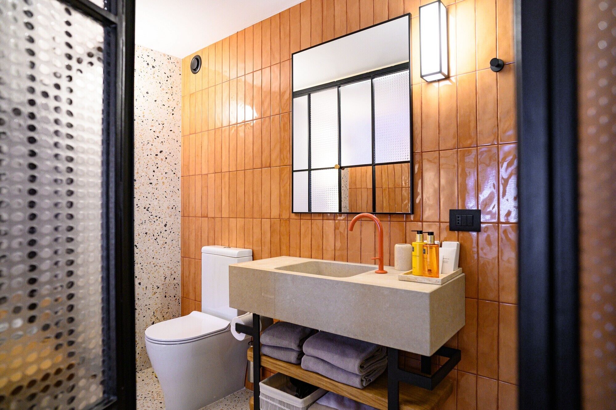 A modern bathroom with a wooden panel wall, a large mirror, a sink with toiletries, and a toilet.