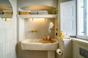Une salle de bain avec un lavabo en pierre, un miroir, un porte-serviettes avec des serviettes, une plante en pot et des articles de toilette rangés sur une étagère.