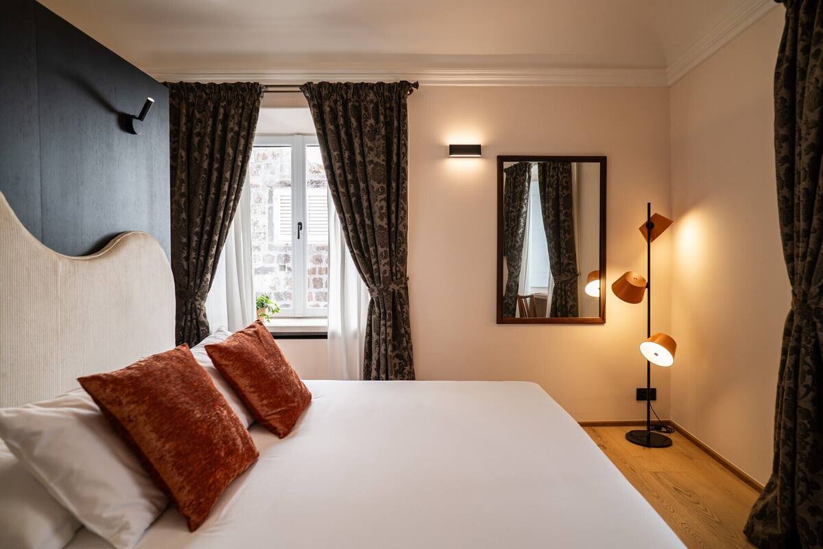 Habitación de hotel con cama, dos almohadas, ventana con cortinas, espejo en la pared y lámpara de pie.