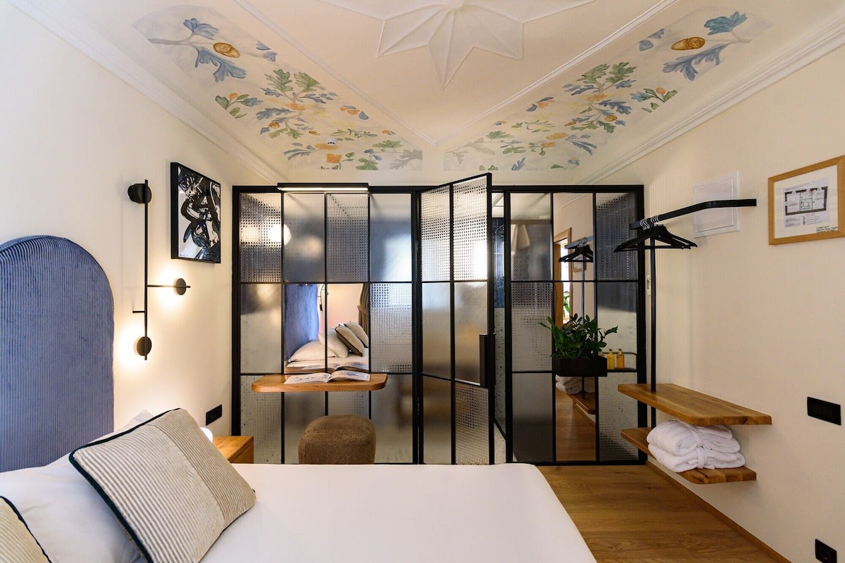 Habitación de hotel pequeña con cama, escritorio, silla, planta y estante con toallas.