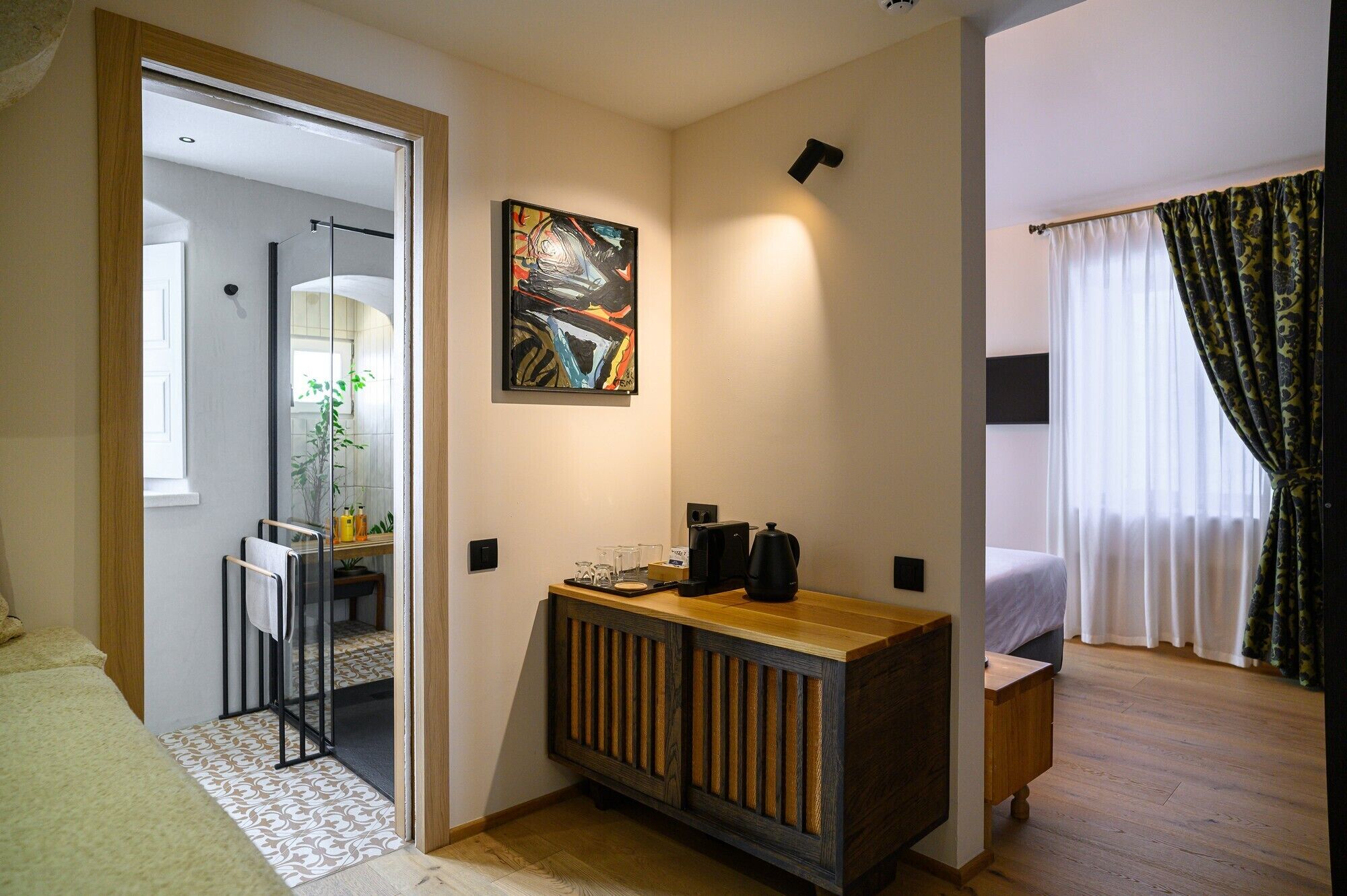 A hotel room with a bed, a bedside table with a coffee maker, a painting on the wall, and a view of the outside through a window with curtains.