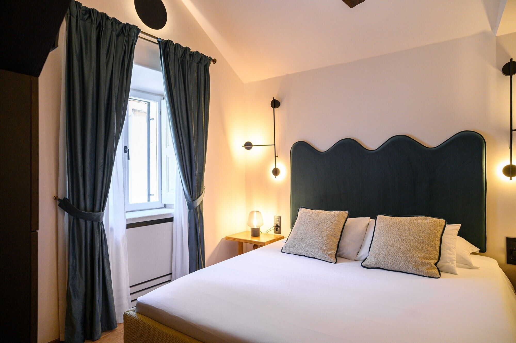 A hotel room with a bed, curtains, a nightstand, a lamp, and a wall-mounted light fixture.