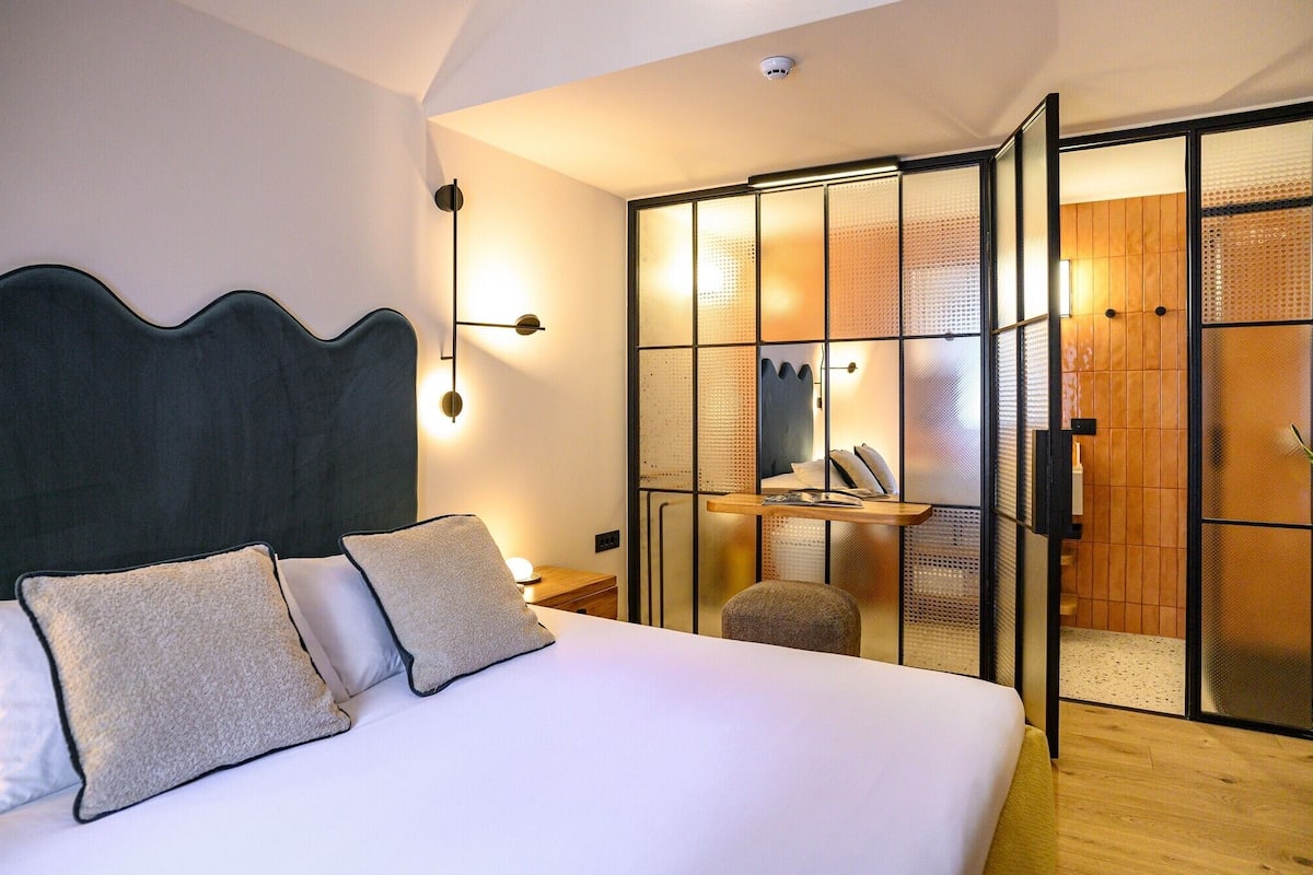 Un dormitorio moderno con una cama grande, una mesita de noche, una lámpara de pared y una puerta corrediza de vidrio que da al baño.
