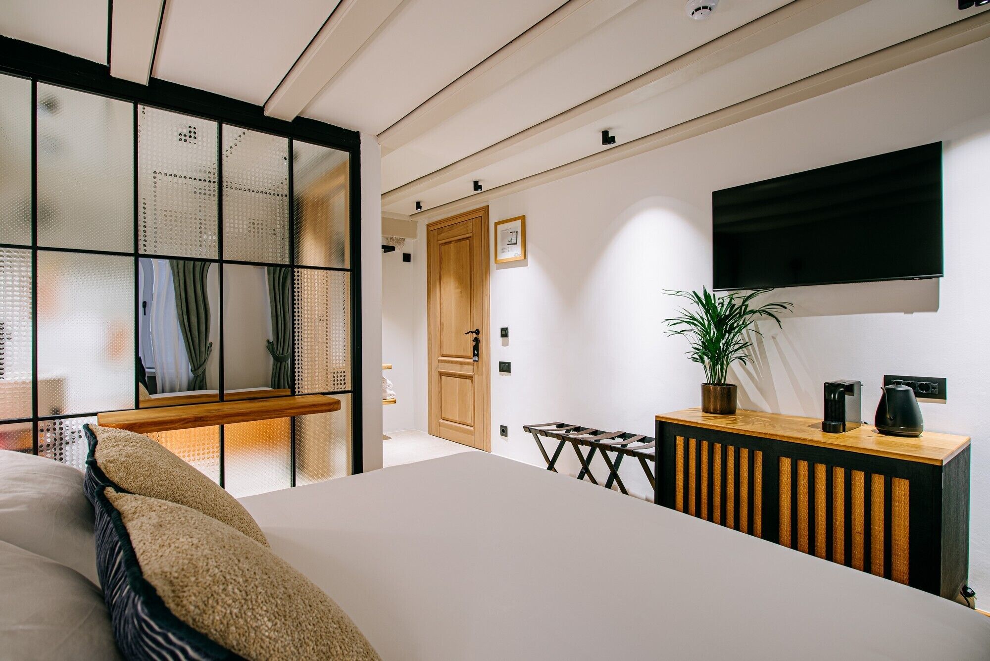 Quarto moderno com cama grande, mesa de cabeceira de madeira, planta em vaso e televisão fixada na parede.