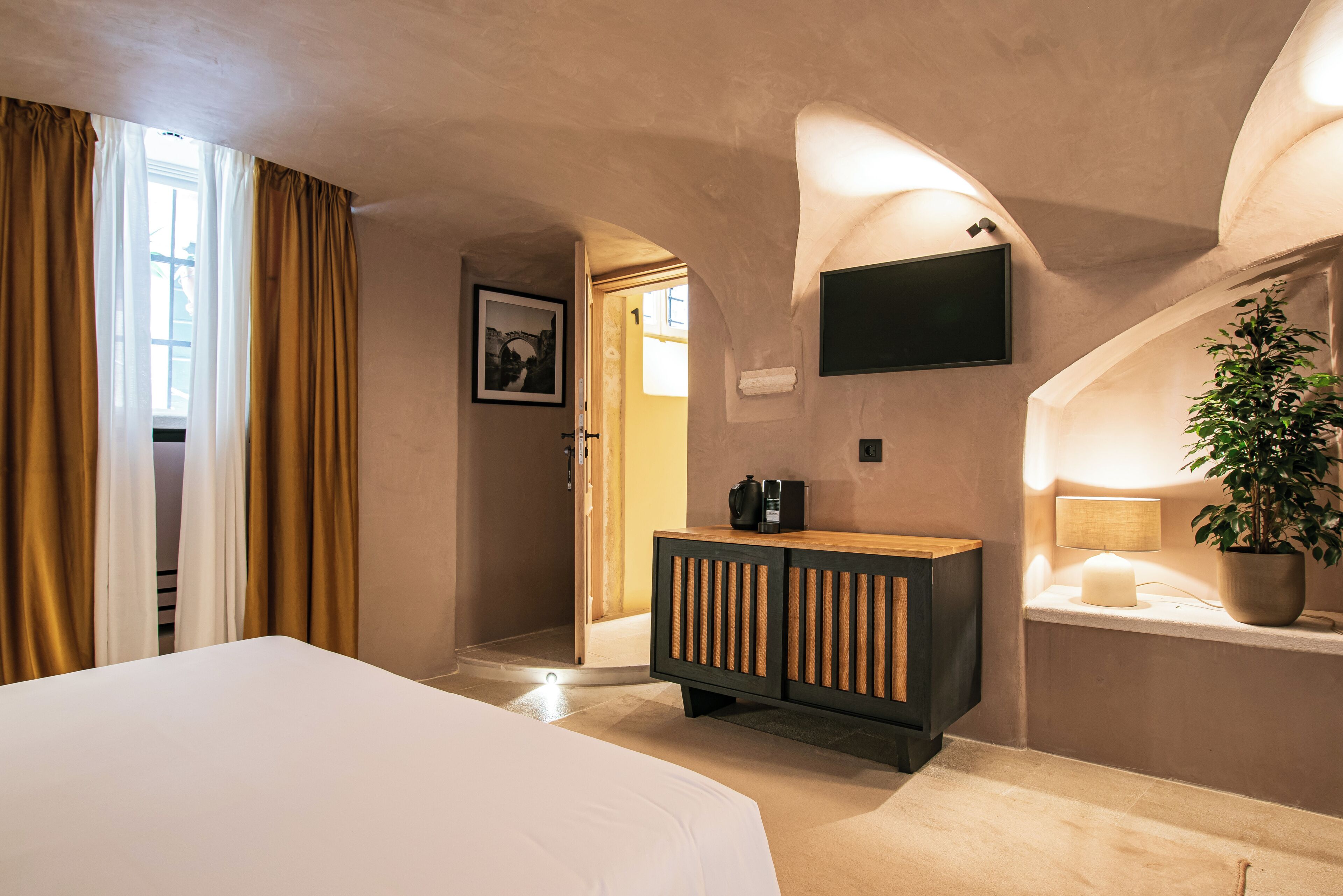 A hotel room with a bed, a wooden cabinet, a TV mounted on the wall, and a potted plant.