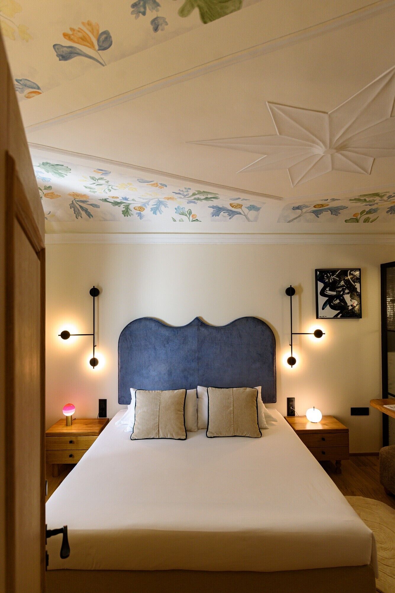 Une chambre à coucher avec un grand lit, deux tables de chevet avec des lampes, un plafond orné de motifs floraux et un tableau abstrait accroché au mur.