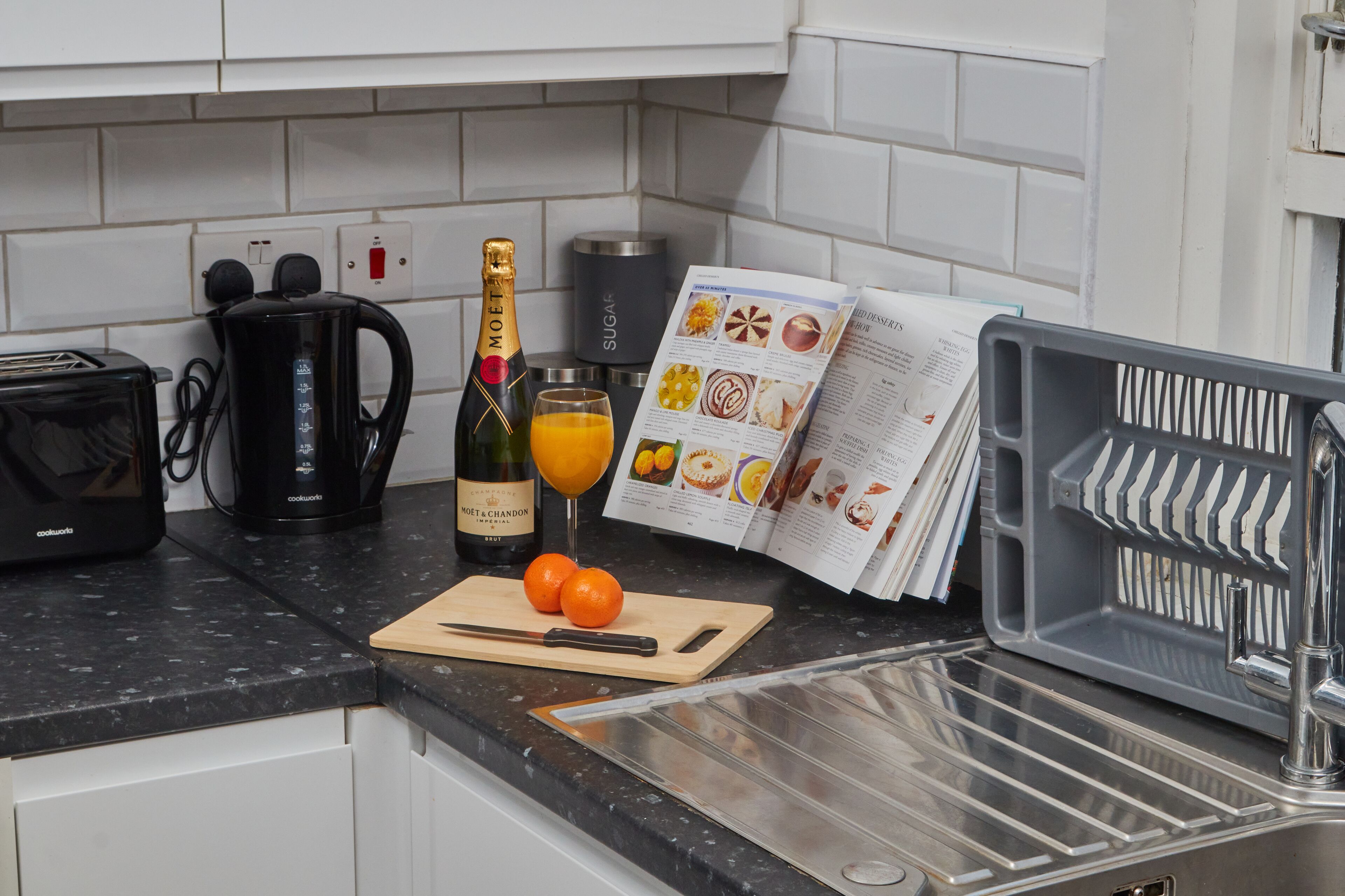 A kitchen counter with a coffee maker, a bottle of champagne, a glass of orange juice, a cutting board with a knife, and a magazine.