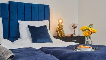 A neatly made bed with blue bedding, a bedside table with a lamp, and a vase with flowers.