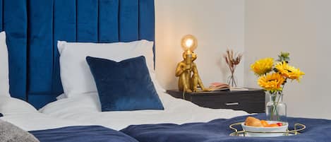A neatly made bed with blue bedding, a bedside table with a lamp, and a vase with flowers.