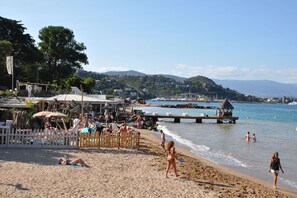 Uma praia com pessoas tomando sol e nadando, um píer e edifícios ao fundo.