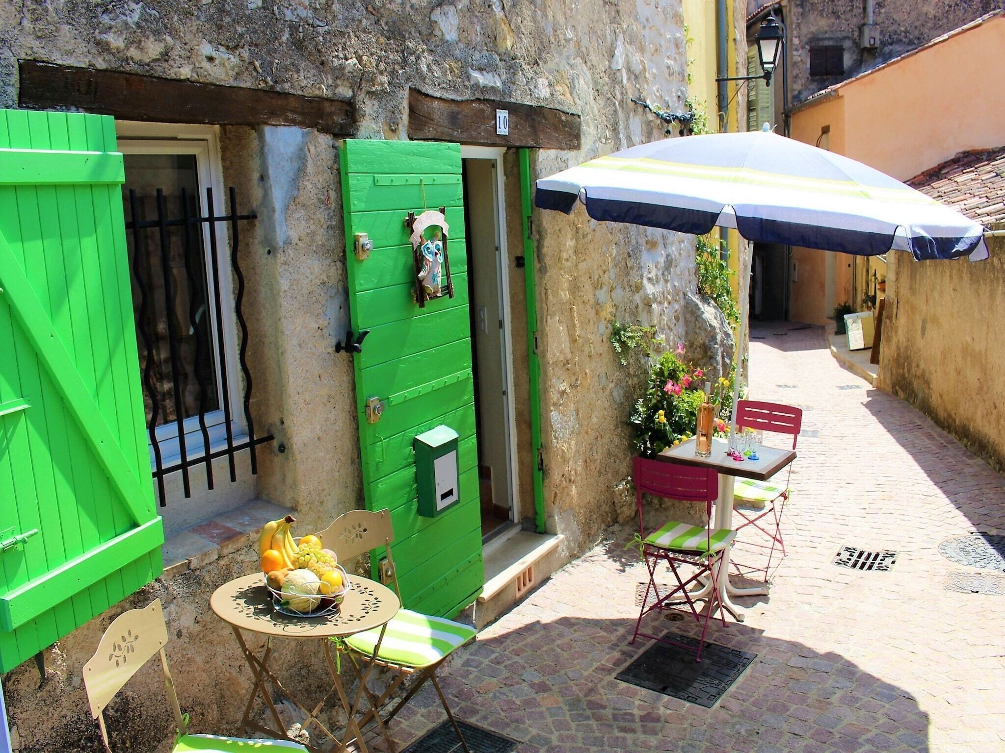 A narrow cobblestone street with a green door, a table with a basket of fruit, and a red chair.