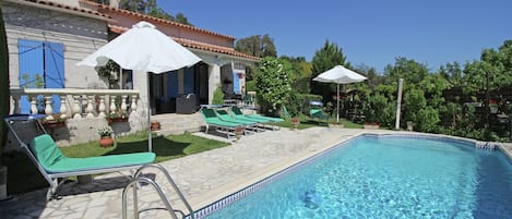 A swimming pool with lounge chairs and umbrellas, surrounded by a stone patio and a building with blue shutters.