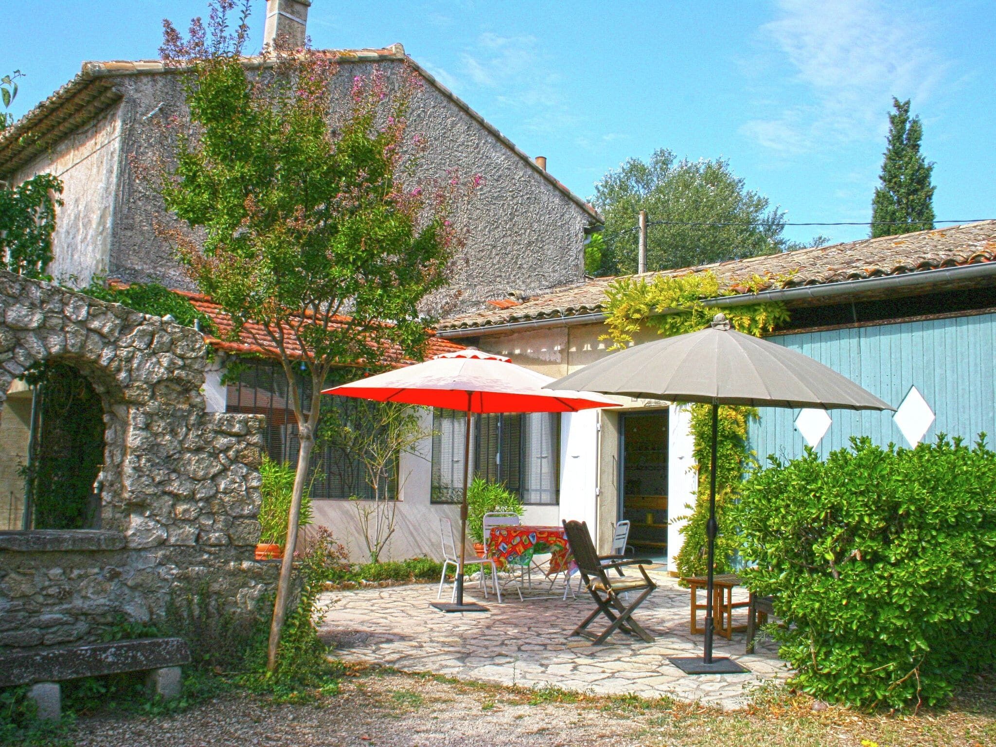 Un bâtiment en pierre avec un toit en tuiles, une terrasse avec une table et des chaises, et un parasol rouge et gris.