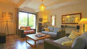 Una sala de estar con un sofá, sillones, una mesa de centro y un televisor. Se aprecia un ventanal con cortinas, una araña y un espejo en la pared.